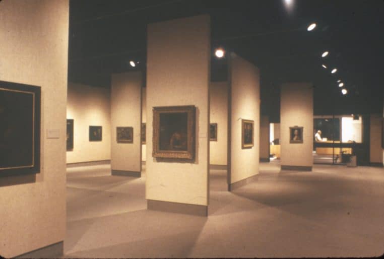 An art gallery, with walls on which paintings are mounted. A figure stands in a reception area.