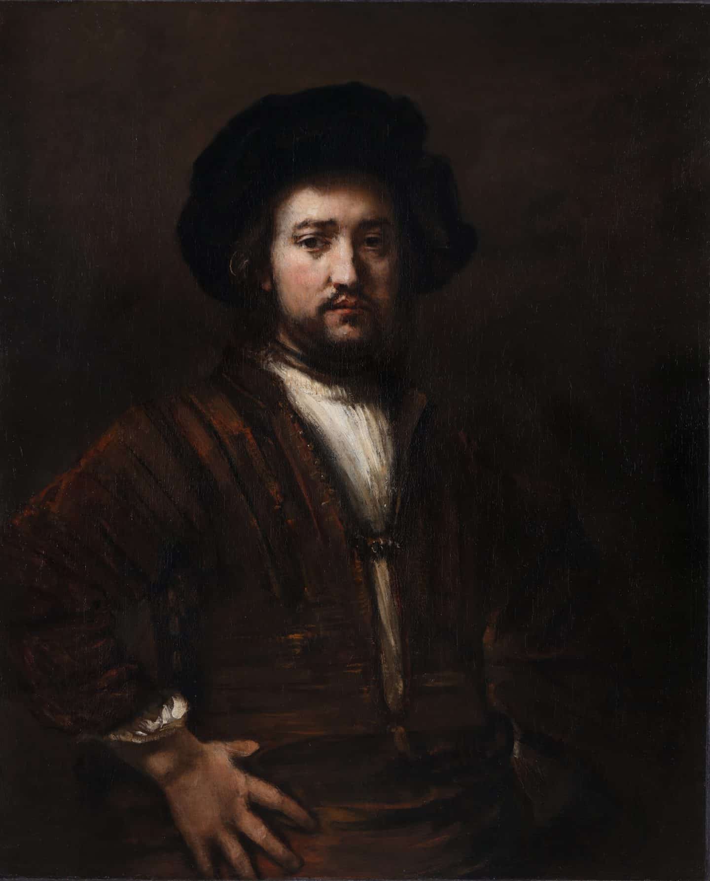 Portrait of a bearded man dressed in a brown jacket over a white shirt and black beret facing us with hands on his hips.