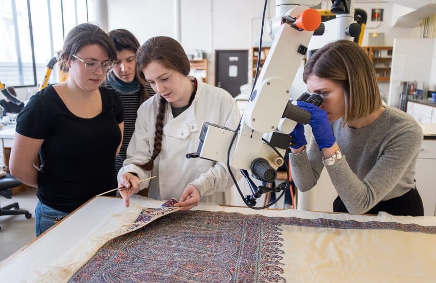 Lorna Rowley speaks with Master of Conservation graduate students while Vanessa Nicholas examines a shawl from the collection through a microscope. Photo: Garrett Elliott