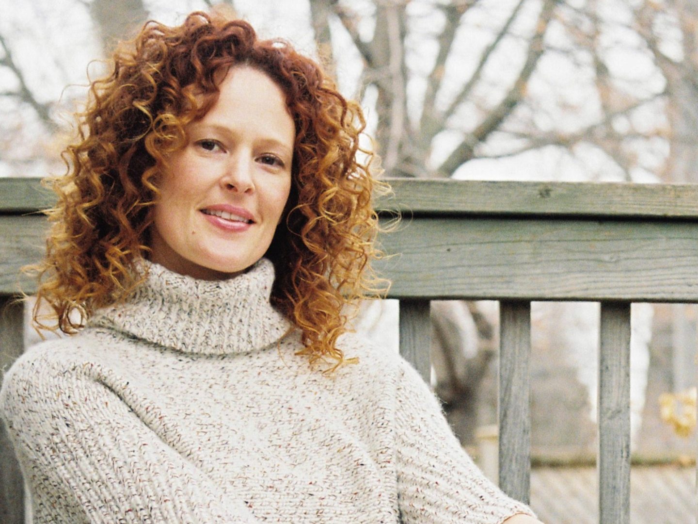 Portrait of Julie Riches, a white woman with curly red hair wearing a light grey knit sweater sitting on an armchair outside.