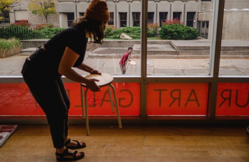 Person holding a stool in front of a glass-paned wall, looking outward to another person. University campus in the background.