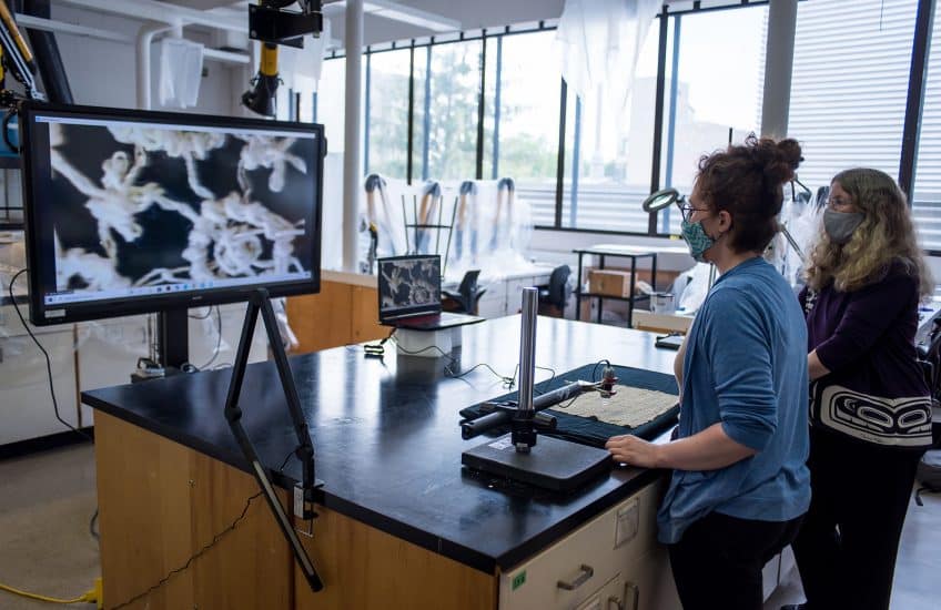 Working in the object conservation lab in Art Conservation's labs , Laura Peers and Anne-Marie Guérin demonstrate the multiple-camera setup used to provide visits with Indigenous ancestors in Agnes’s care.