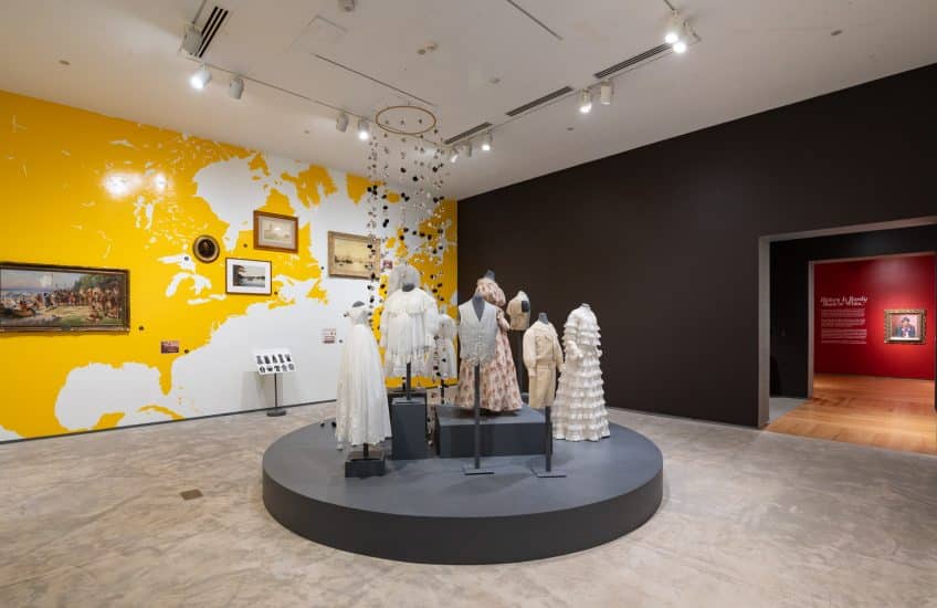 Installation view of History Is Rarely Black or White, showing a yellow vinyl map on an entire wall with artworks hung at the approximate location they depict. In the centre of the room, there is a round grey plinth with a display of cotton garments, such as dresses, vests and children's suits.