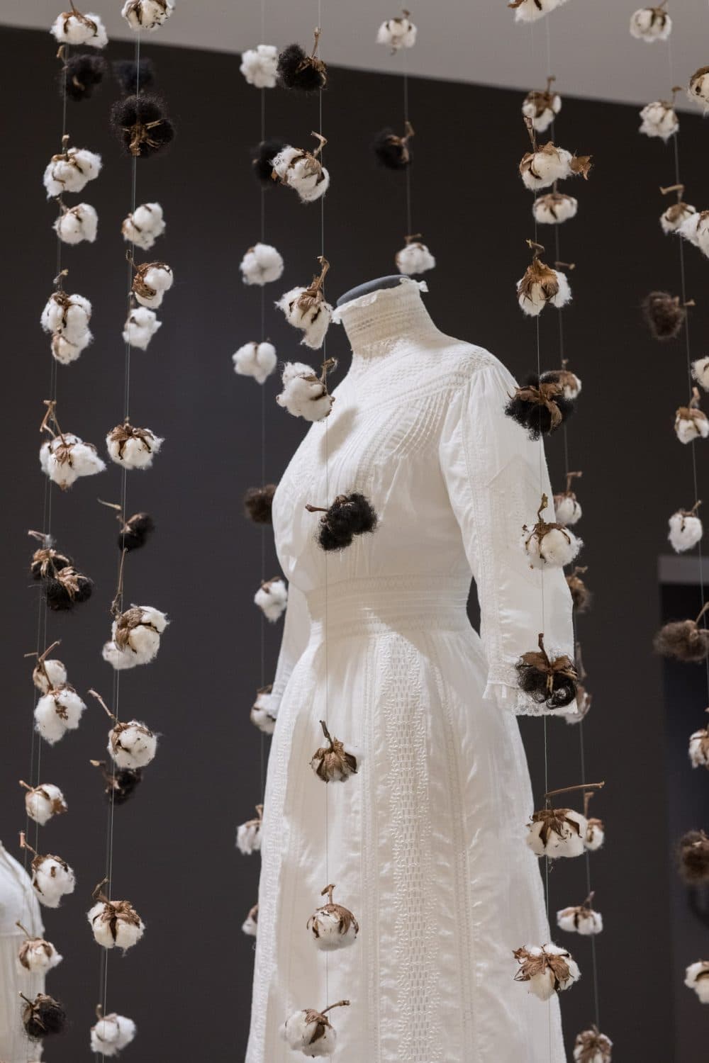 Karin Jones’ Freed installation featuring strings of raw cotton bulbs and Black hair suspended around a wedding dress from 1893 from the Collection of Canadian Dress.