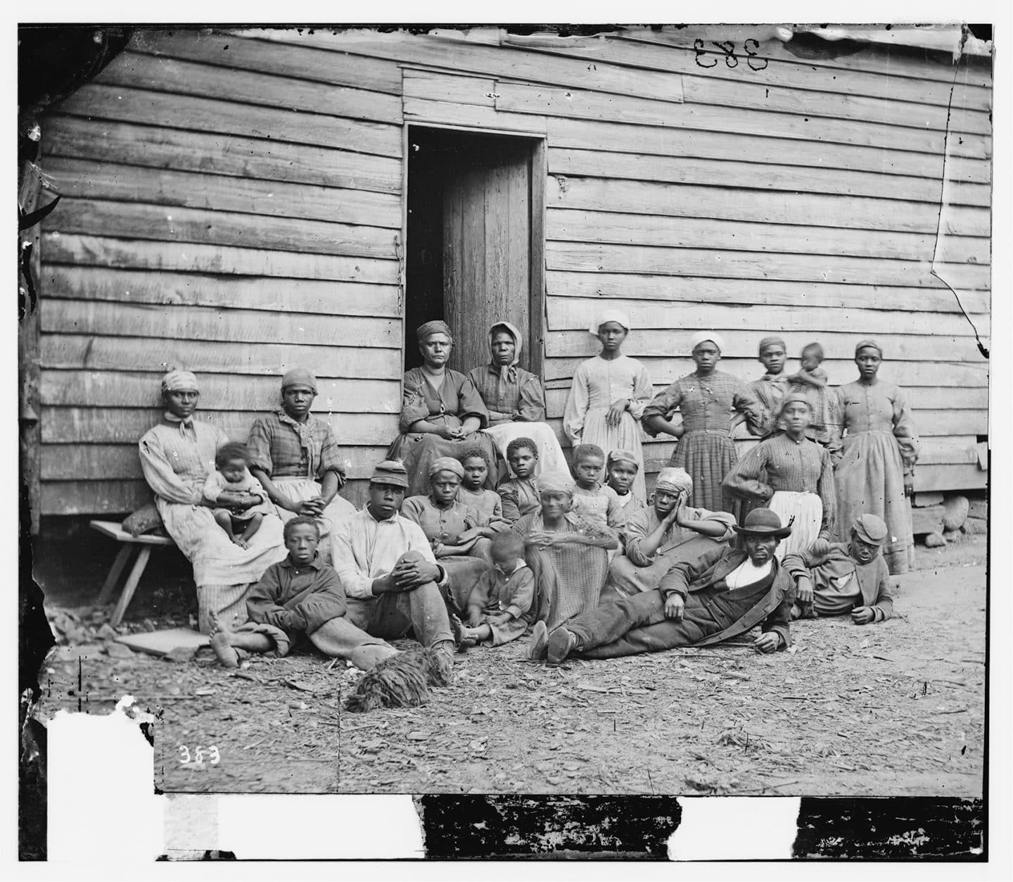 An archival image of a group of Black people sitting outside a wooden house from 1862.