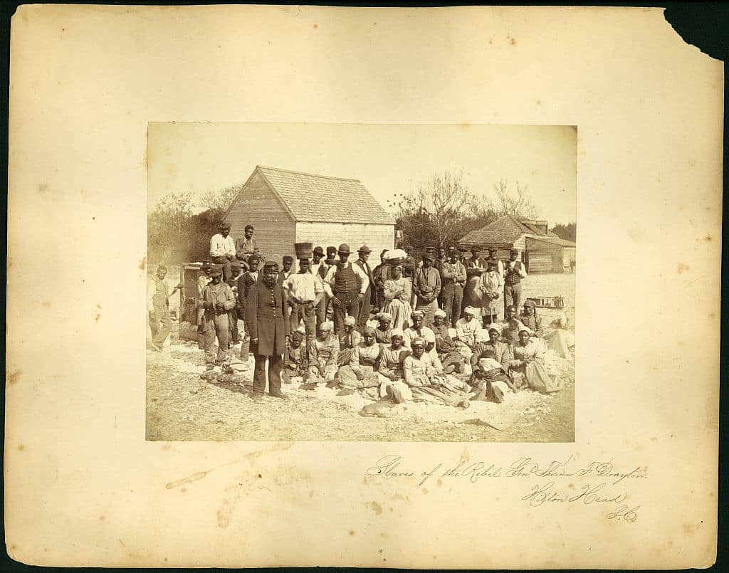 An archival image of a group of Black people sitting and standing. The image is inscribed: "Slaves of the rebel Genl. Thomas F. Drayton, Hilton Head, South Carolina, 1862"
