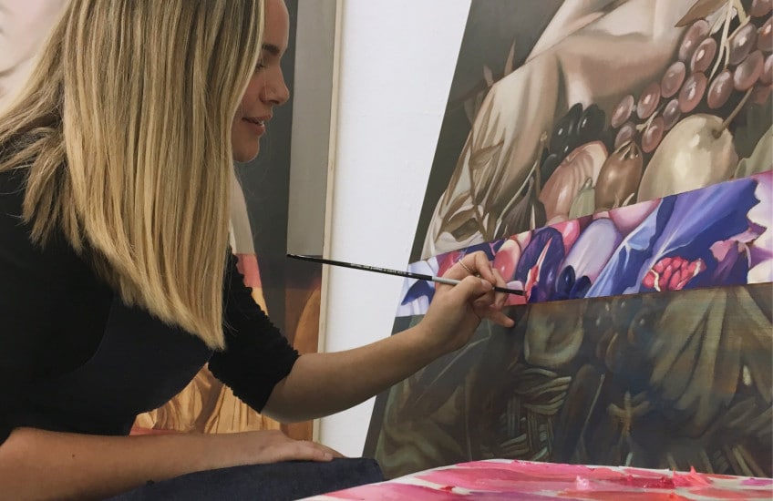 Portrait of Emily Joyce painting a large canvas. She is wearing black and is turned to the side with the paint brush in her left hand.