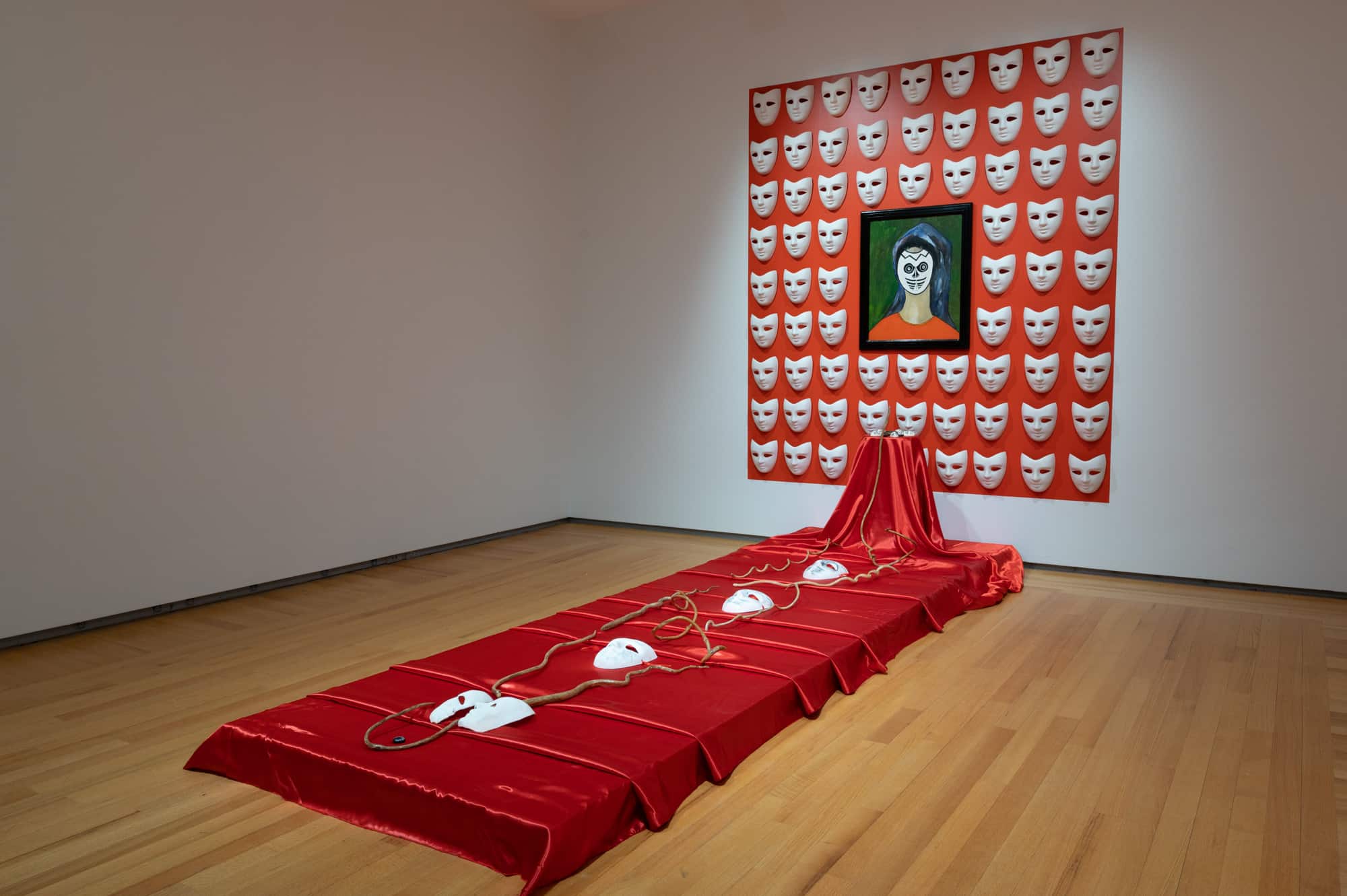 Installation image of Winsom Winsom's The Masks We Wear, 2018. Shows a long, low plinth on the floor covered in red satin fabric upon which are white masks entwined with cord. There is a small altar that the cord reaches up to. Behind this is a red square on the wall which is superimposed with rows of the white masks. In the middle is a painting of a figure wearing a mask in a black frame.