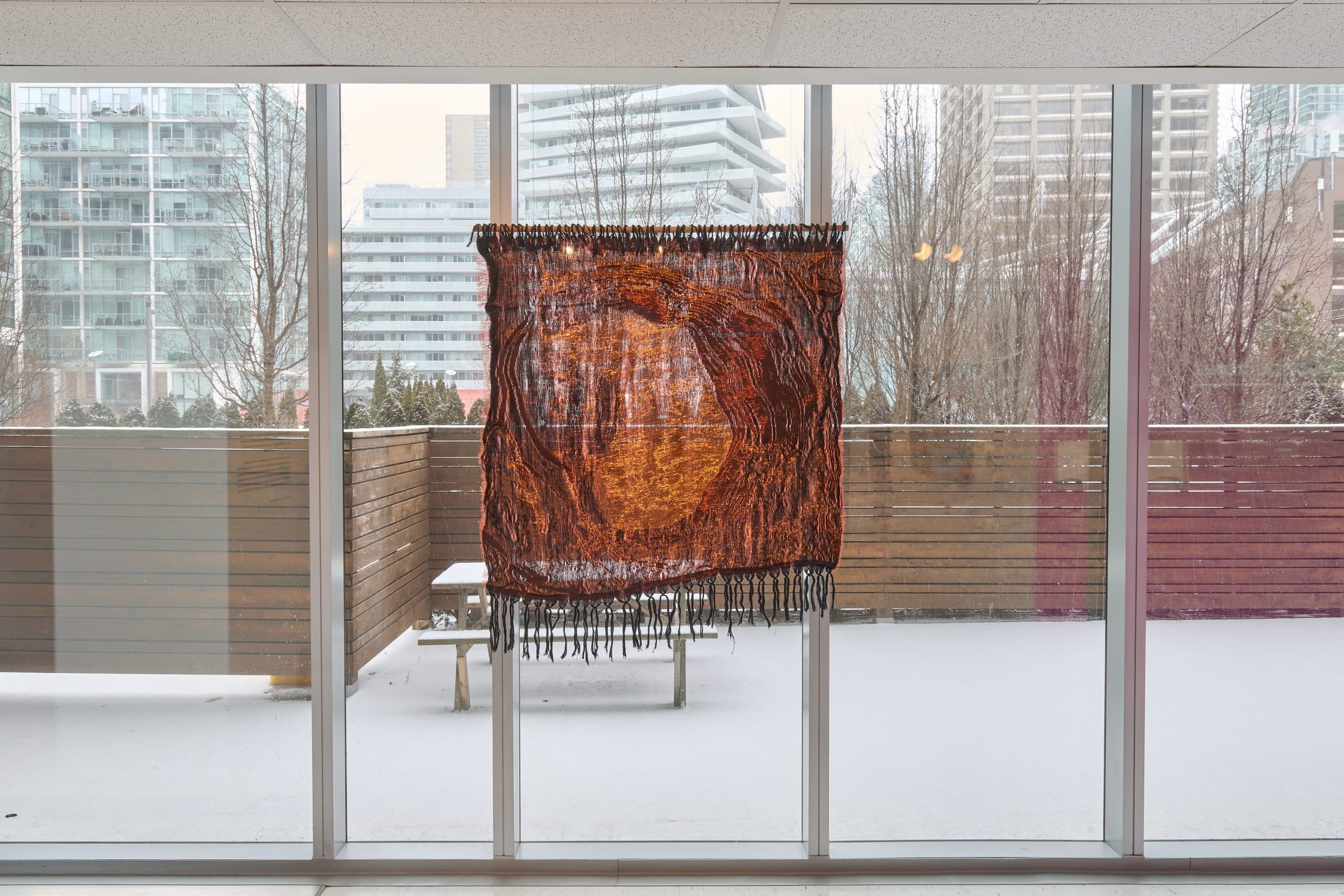 A square shaped shimmering, copper coloured semitransparent woven artwork hanging in front of a bright window.