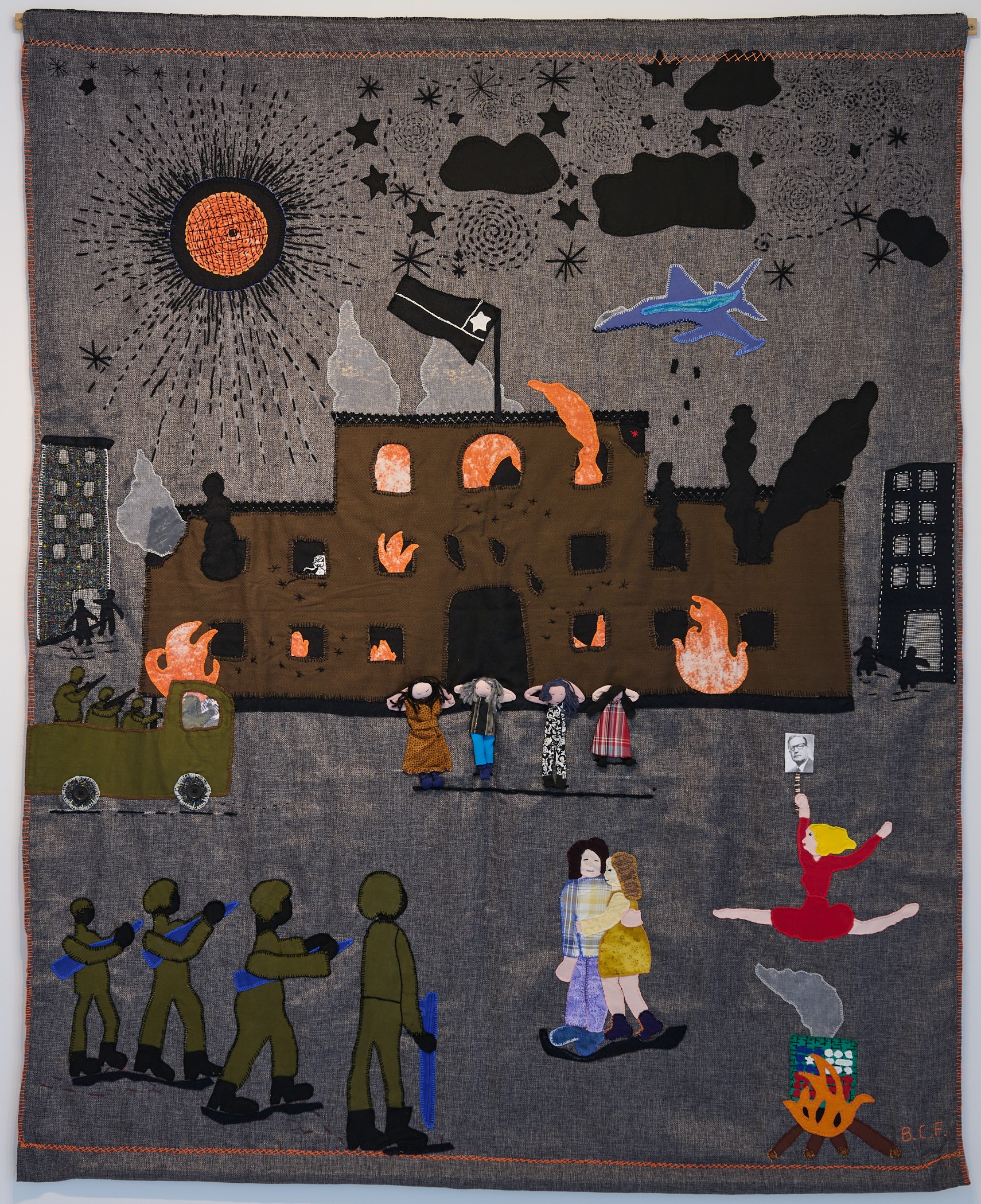 A dark arpillera depicting a violent scene with a burning building, military vehicles, soldiers with guns and women being held at gunpoint.