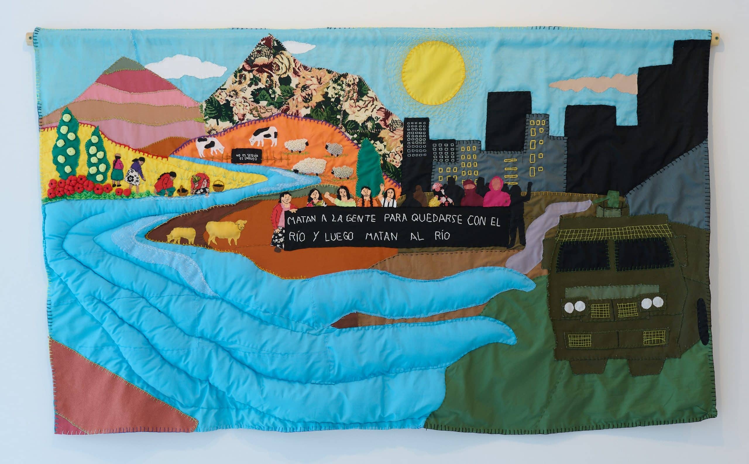 An arpillera depicting a scene with colourful mountains and fields, a blue river, a city, military vehicle and individuals holding a protest sign.