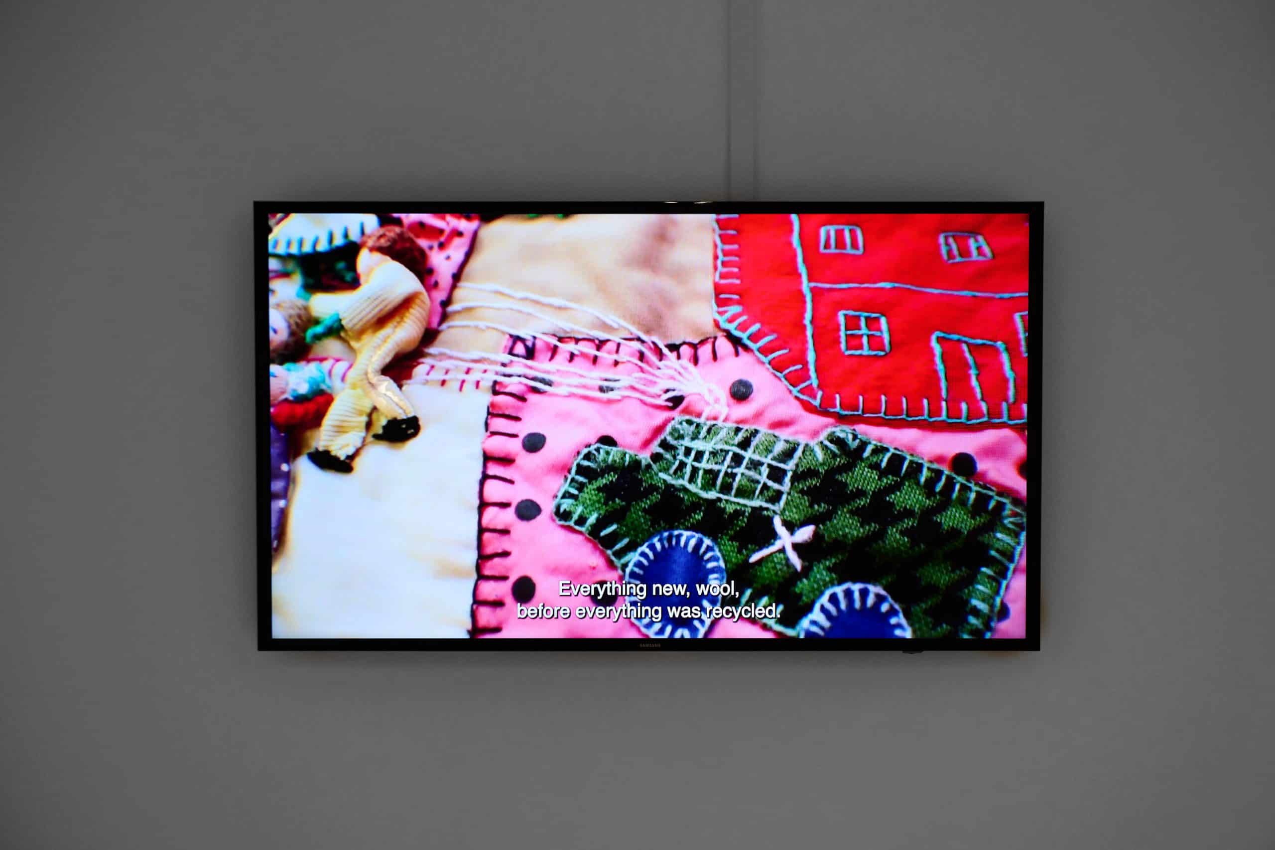 A TV screen hung on a white wall. On screen is a close up of bright pink, red and green patchwork fabrics and blanket stitching. Captions read “Everything new, wool, before everything was recycled.”