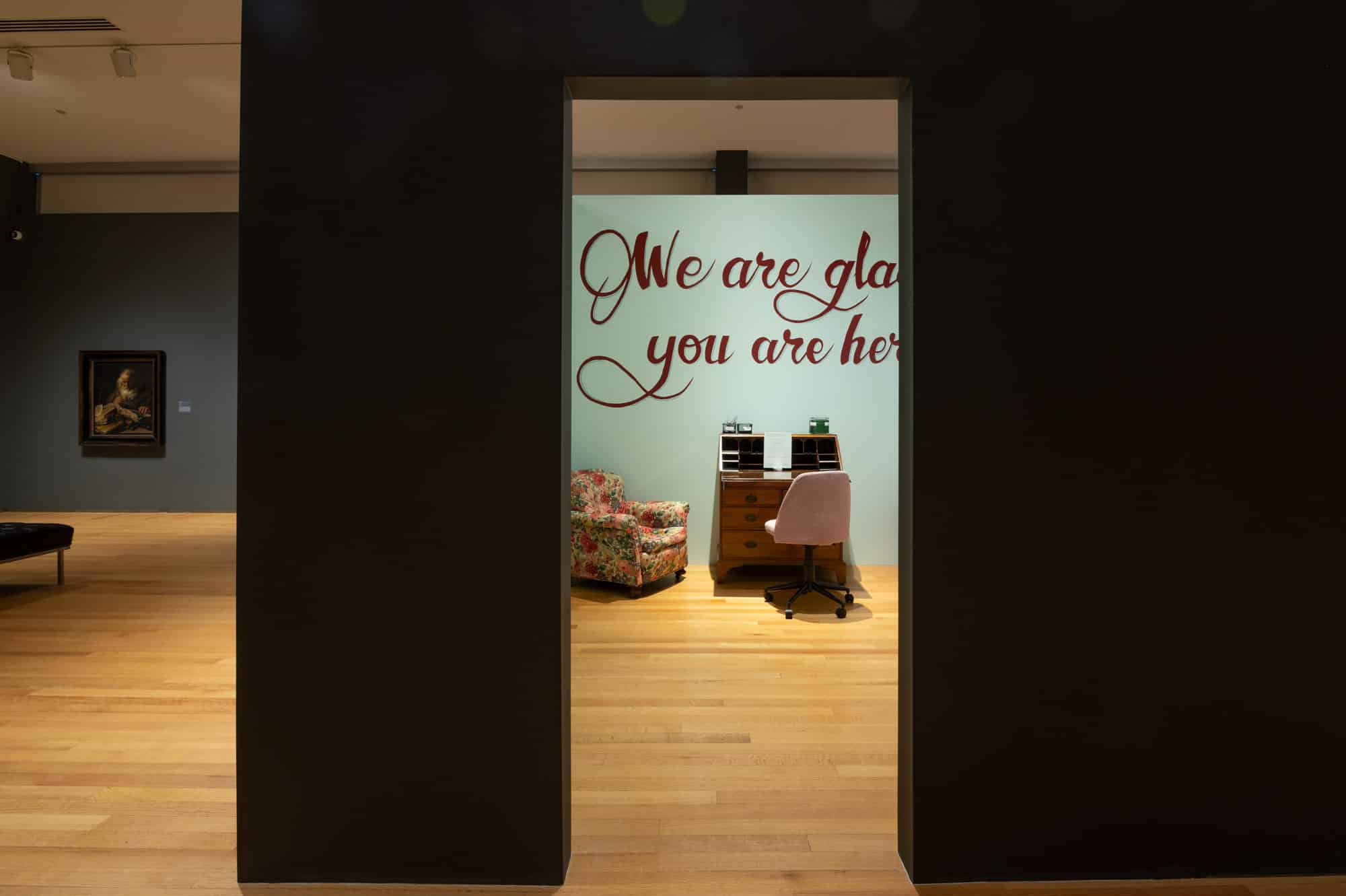 An art gallery showing a desk with a desk chair, an armchair, and a wall with the text “We are glad you are here”.