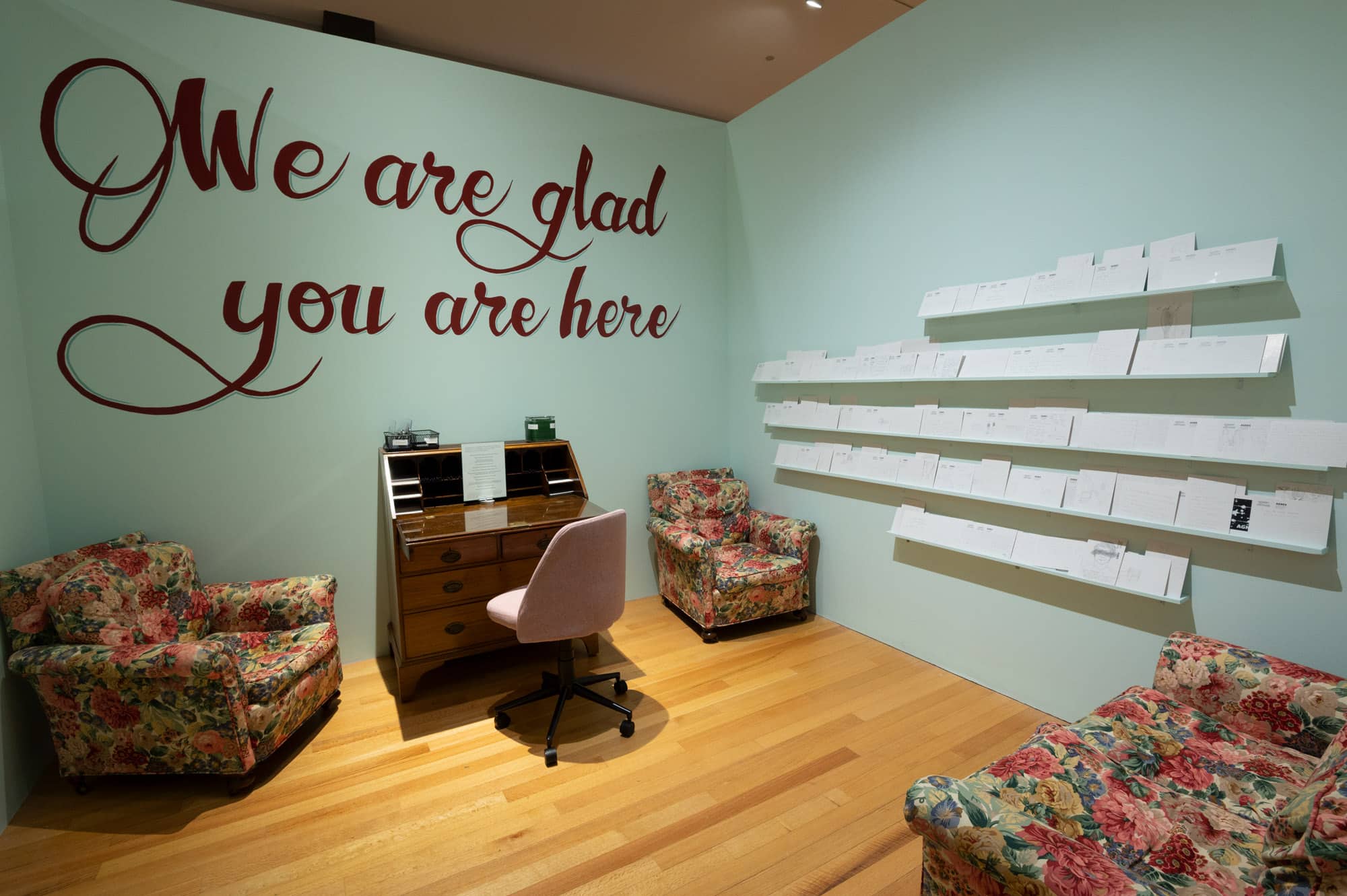 A room with a desk and desk chair, two armchairs, and a love seat. The text “We are glad you are here” is written above the desk.