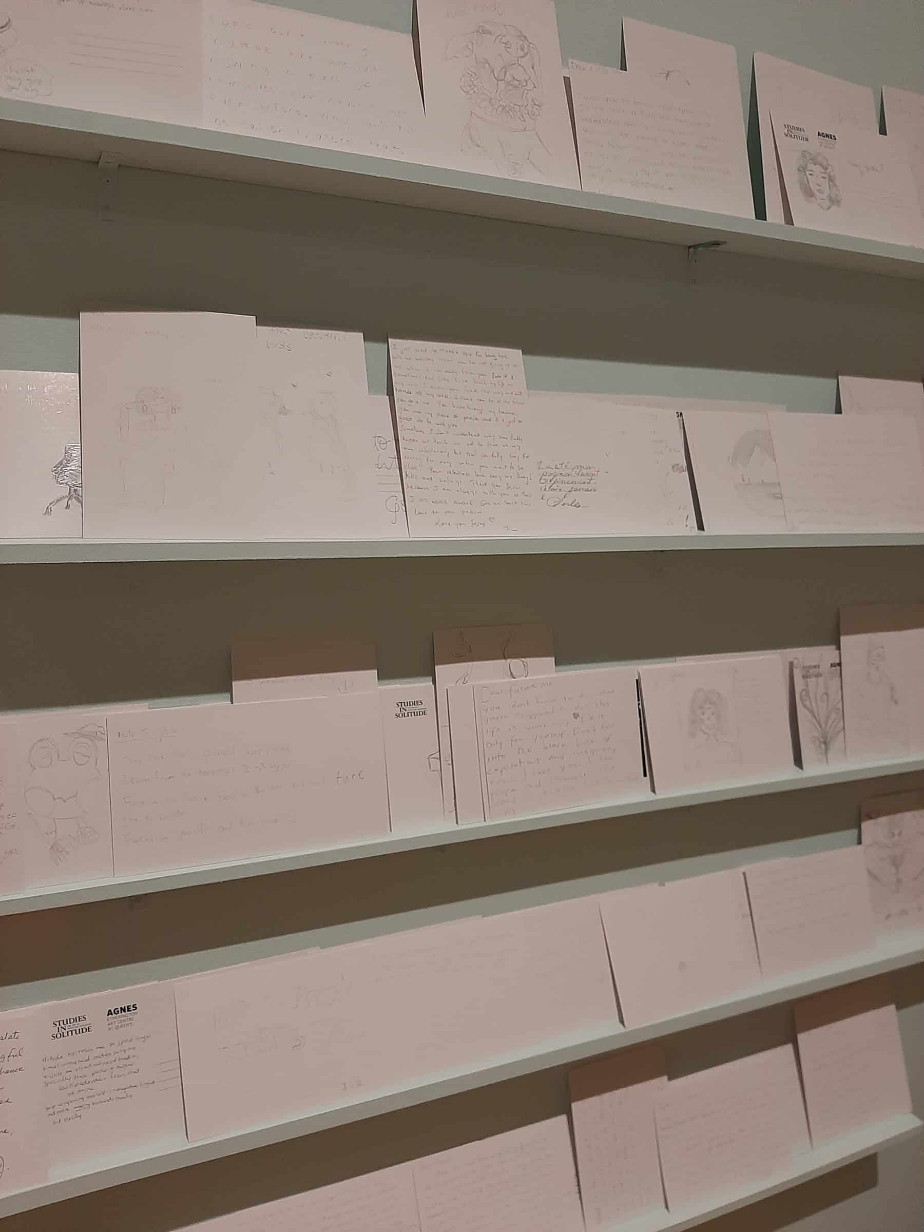 View of shelves with white handwritten postcards, some with pencil drawings, against a light blue wall.