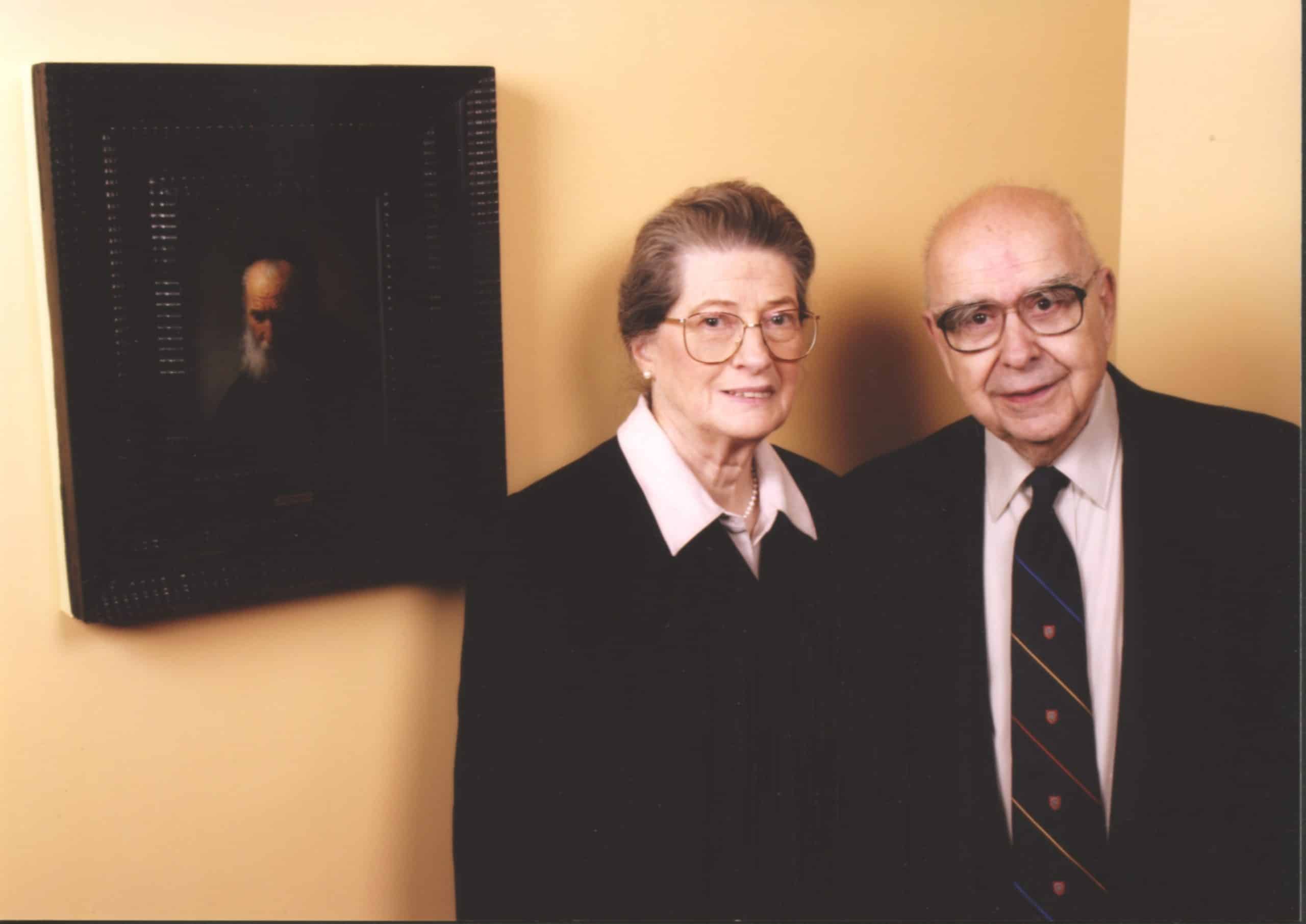 An elderly man and woman wearing glasses and dark clothing stand next to a painting of an old man in a black frame.