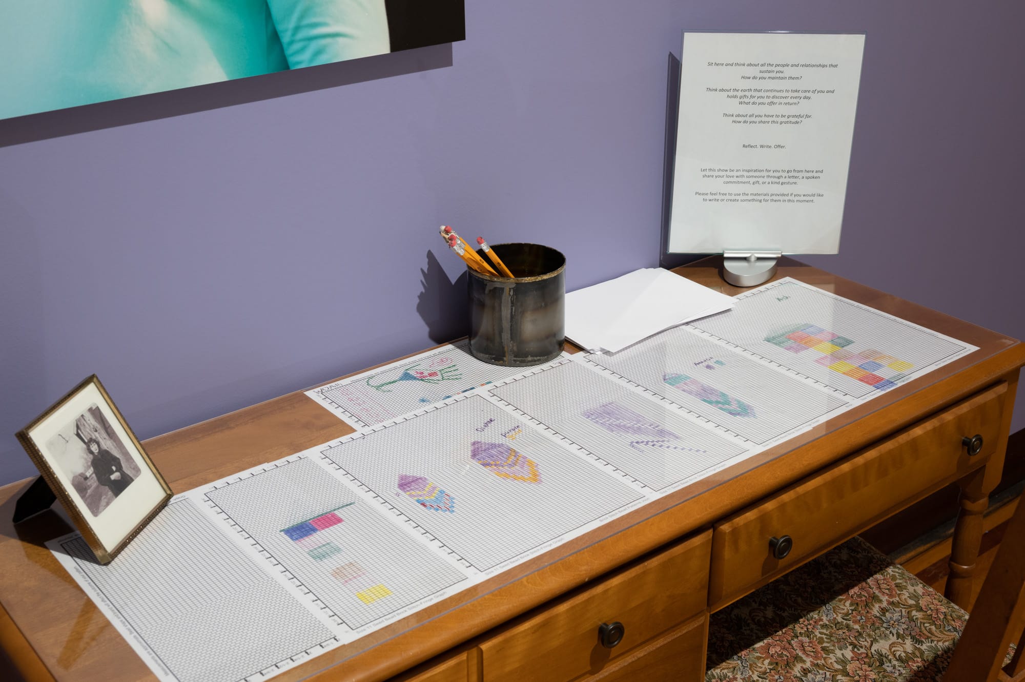 A desk with design sheets for beaded earrings under Plexiglas. A cup of pencils and paper are on the desk.