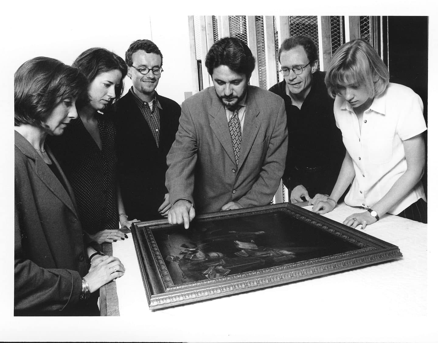 A professor with five students huddle around a painting on the table in front of them.