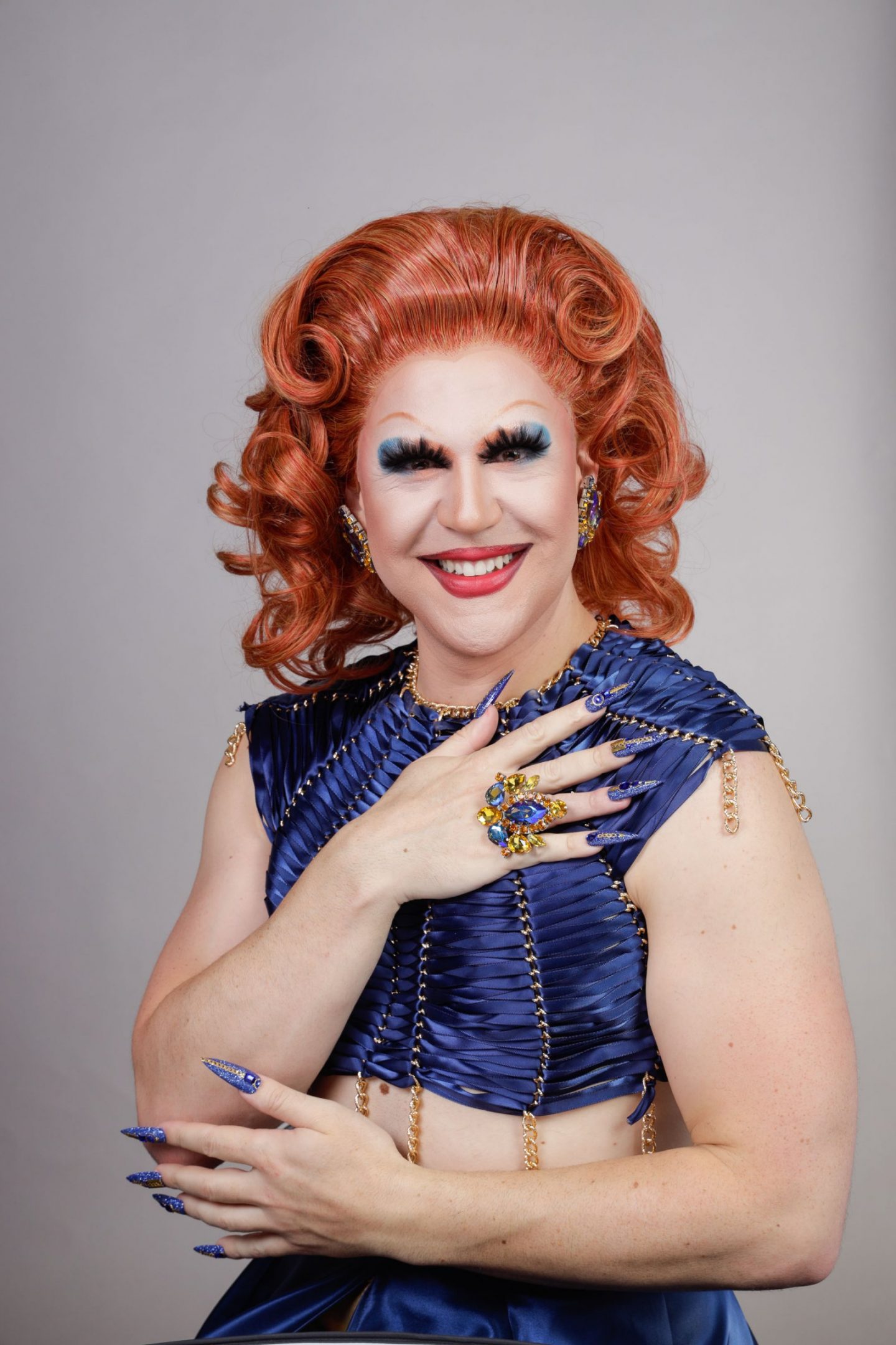 Portrait of drag performer Rowena Whey. She has orange coiffed hair, wears a navy blue dress. She wears long nails and a large ring, matching her dress.