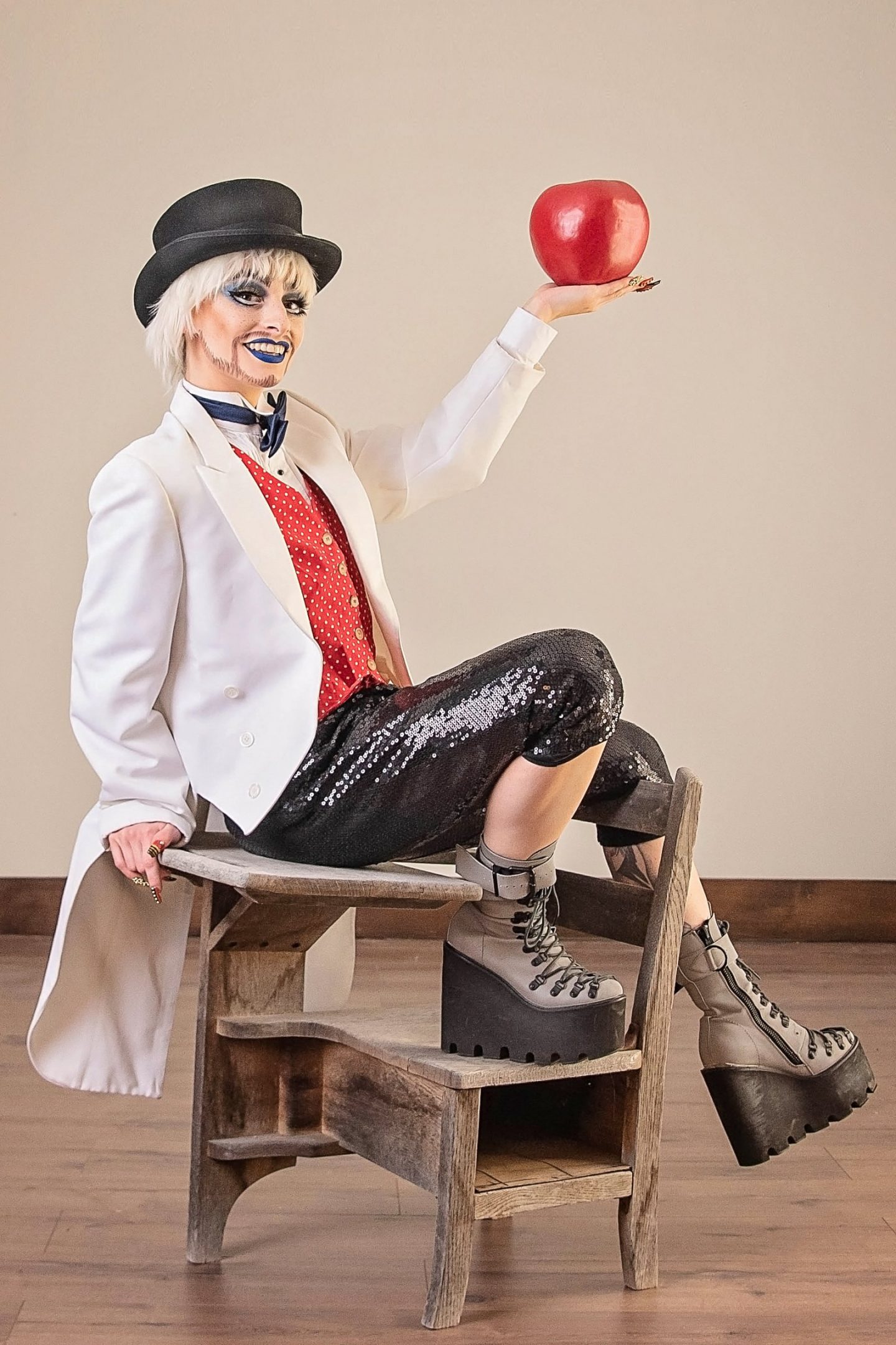 Dare de LaFemme sits jauntily on a small wooden desk holding a large red apple. They are wearing a black hat, white waistcoast, red polka dot vest with a blue bowtie, and black sequin capris pants.