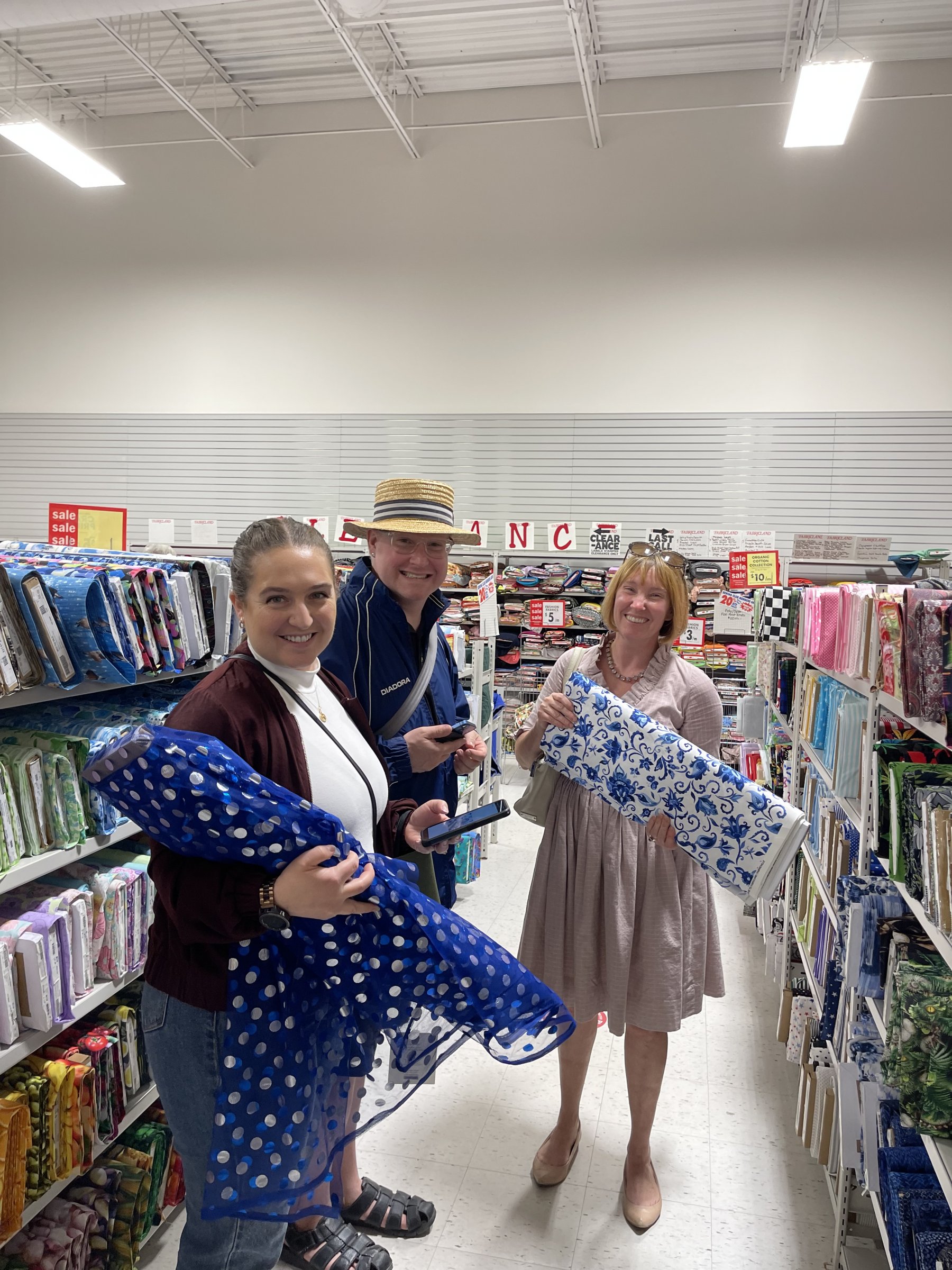 At the store, Marissa Monette holds blue polka dot tulle, Carolyn Dowdell holds a blue delft patterned fabric. Tyffanie Morgan smiles in the middle.