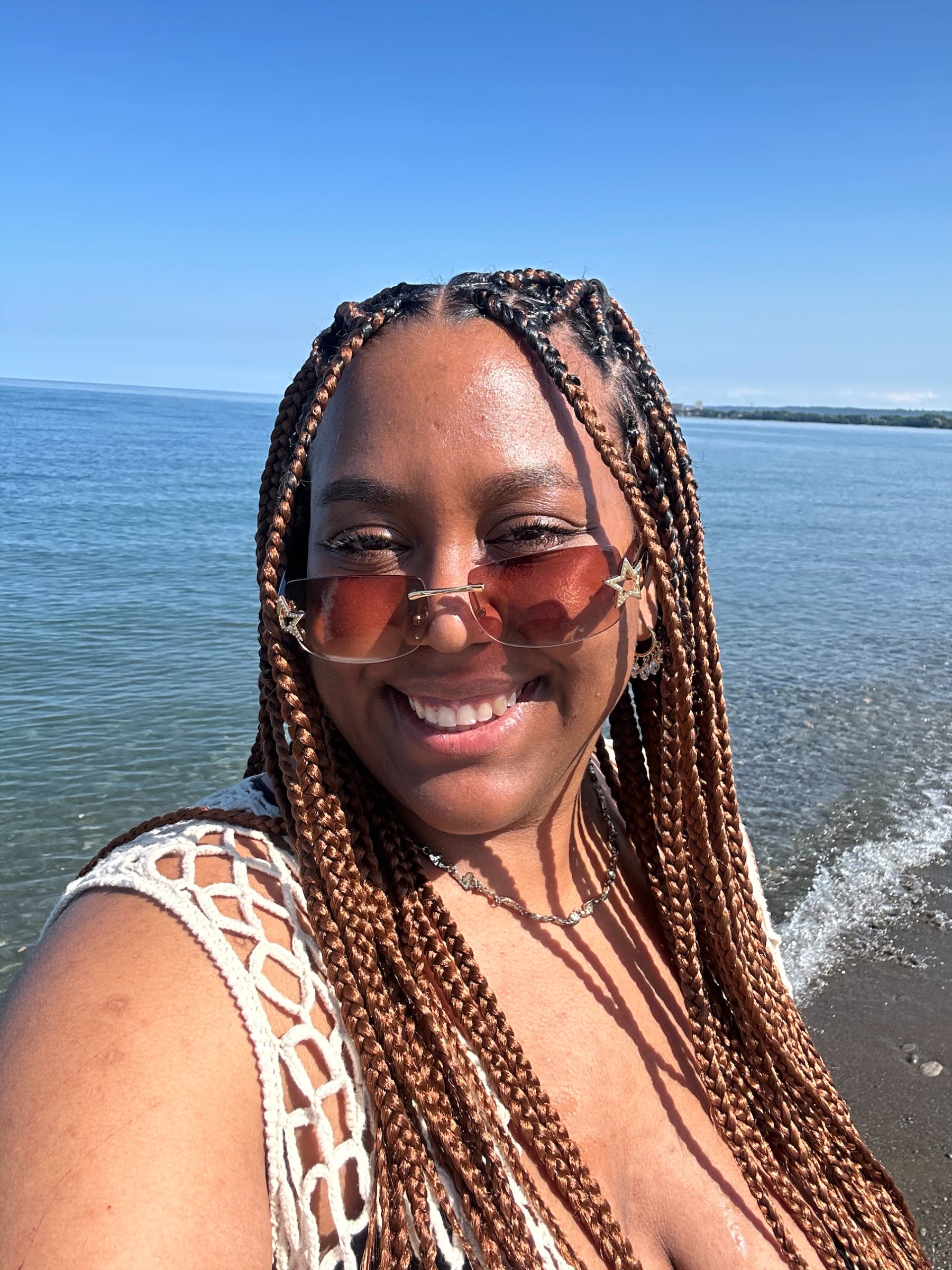 Portrait of Alyssa Vernon standing in front of a body of water. They are wearing sun glasses and have a smile on their face.