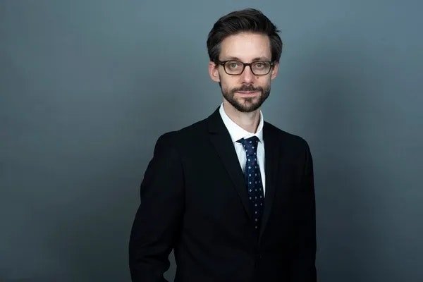 Portrait of Patrick Staheli, a white man with short brown hair, dark rimmed glasses and a beard. He is wearing a dark suit and a blue tie with small polka dots.