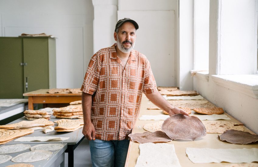 Sameer standing among his flatbreads, looking towards the camera, holding up a flatbread