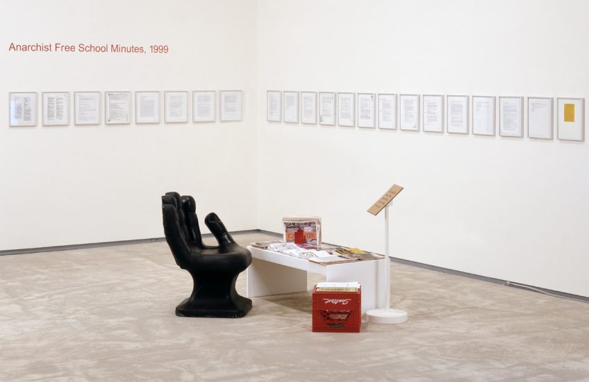 Installation view of Luis Jacob, Anarchist Free School Minutes, 1999, (2002) showing a black hand chair, a low table, a red milk crate, and pages of the manifesto framed on the wall in the corner of a room.