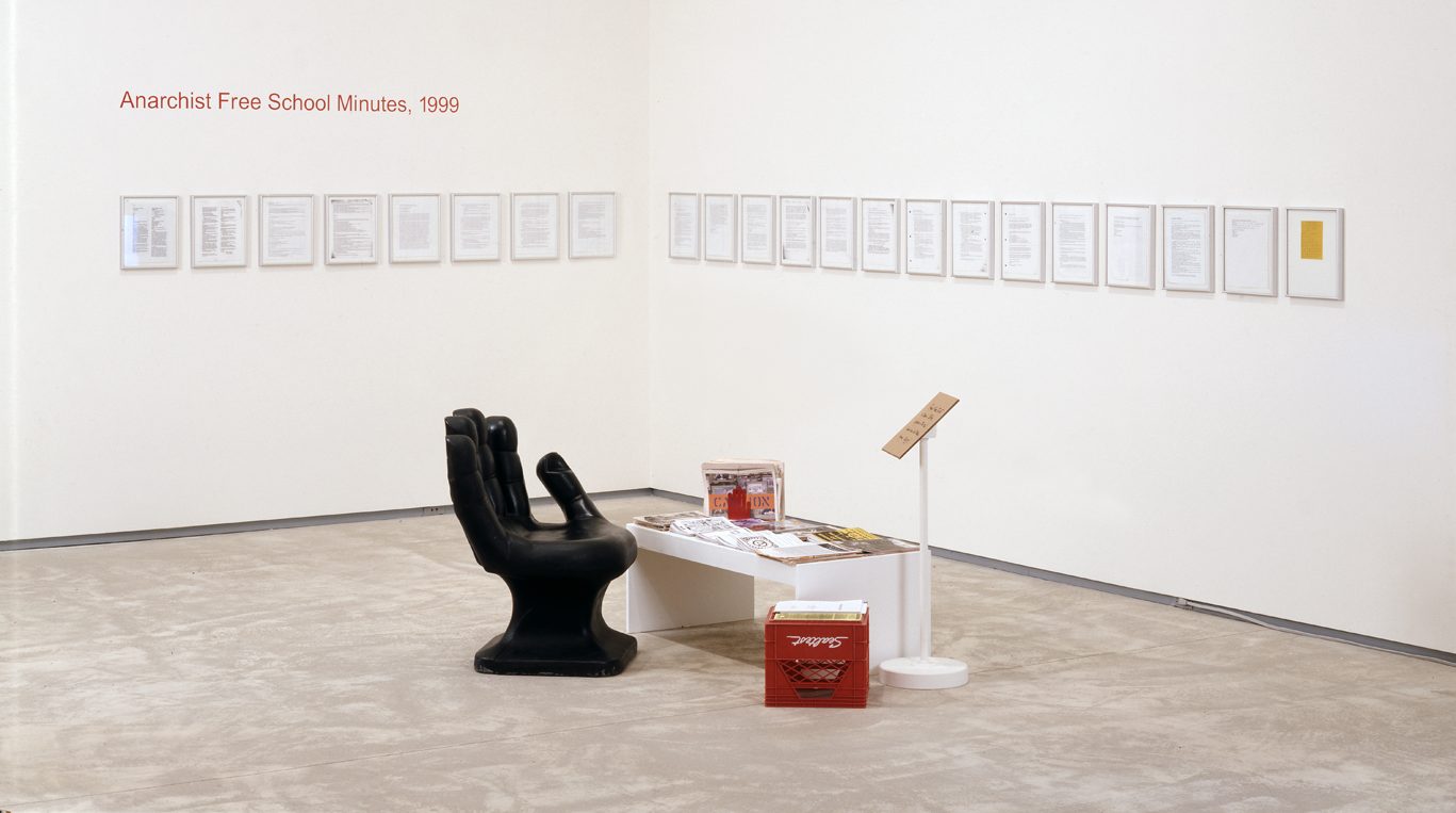 Installation view of Luis Jacob, Anarchist Free School Minutes, 1999, (2002) showing a black hand chair, a low table, a red milk crate, and pages of the manifesto framed on the wall in the corner of a room.