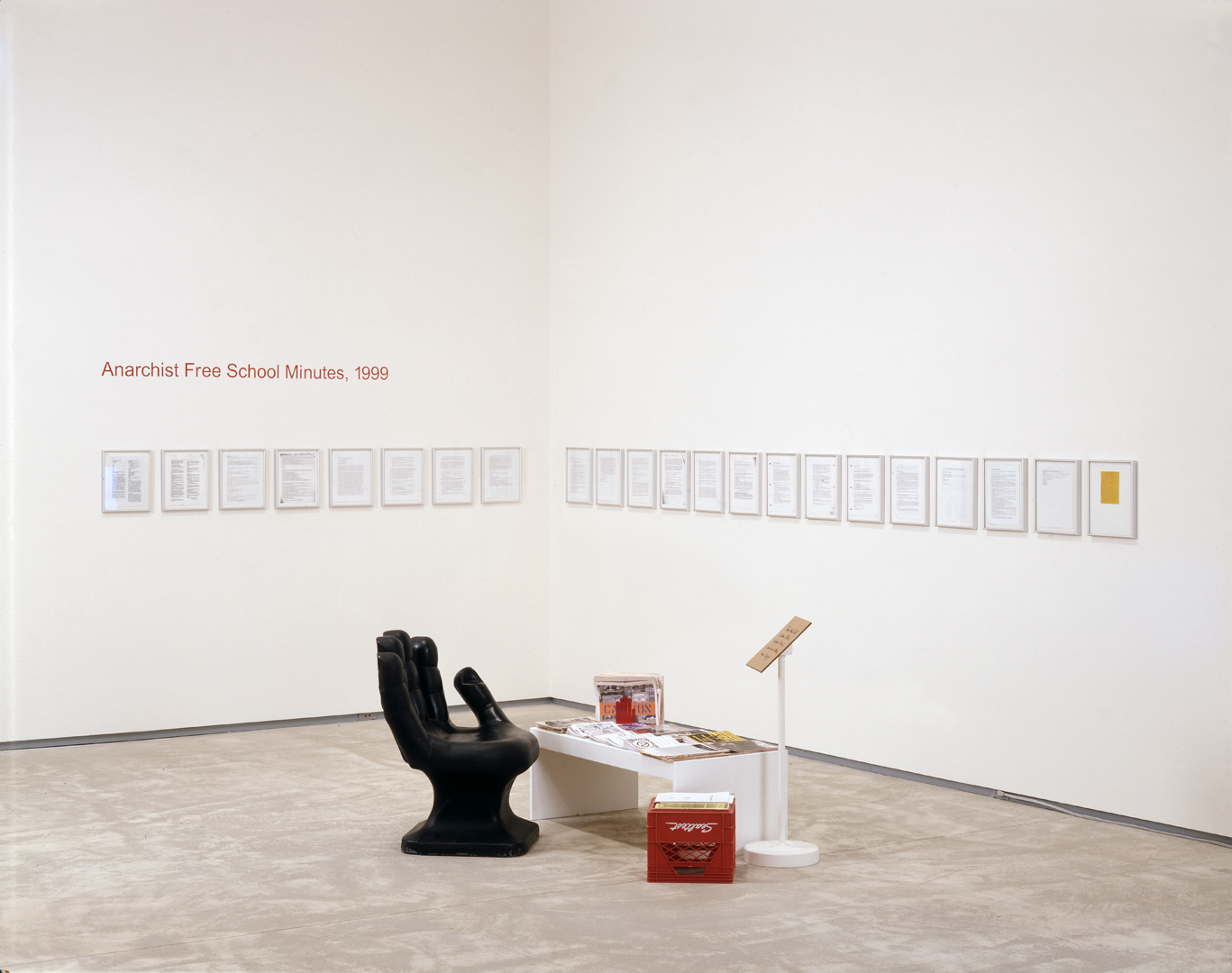 Installation view of Luis Jacob, Anarchist Free School Minutes, 1999, (2002) showing a black hand chair, a low table, a red milk crate, and pages of the manifesto framed on the wall in the corner of a room.