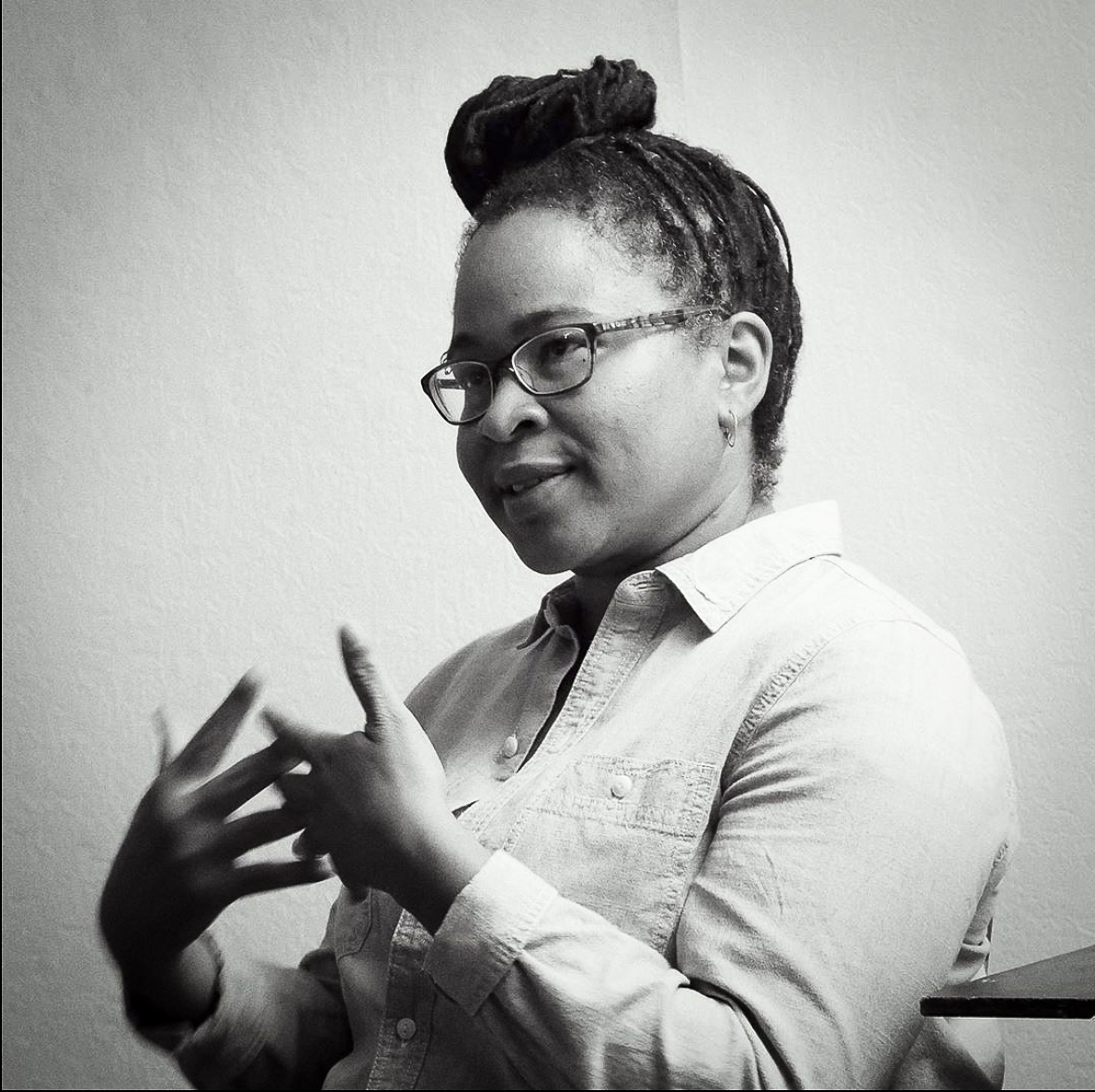 A Black woman is seated, looking to the camera left, explaining something and gesticulating with her hands.