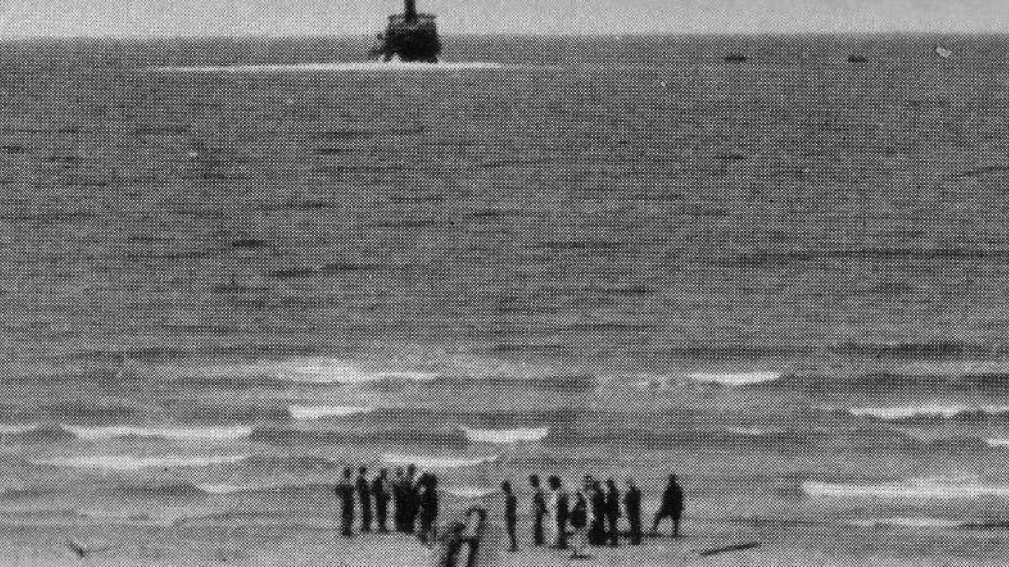 Grainy film still of a coast line with figures of people standing on the shore in the foreground and a boat in the background