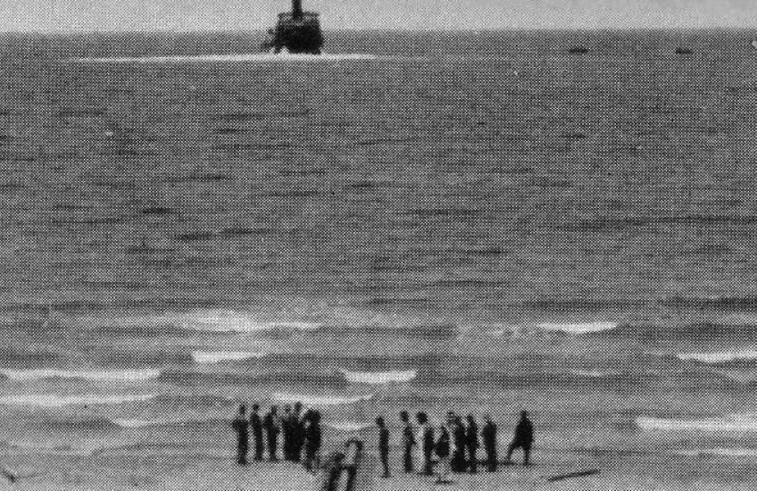 Grainy film still of a coast line with figures of people standing on the shore in the foreground and a boat in the background