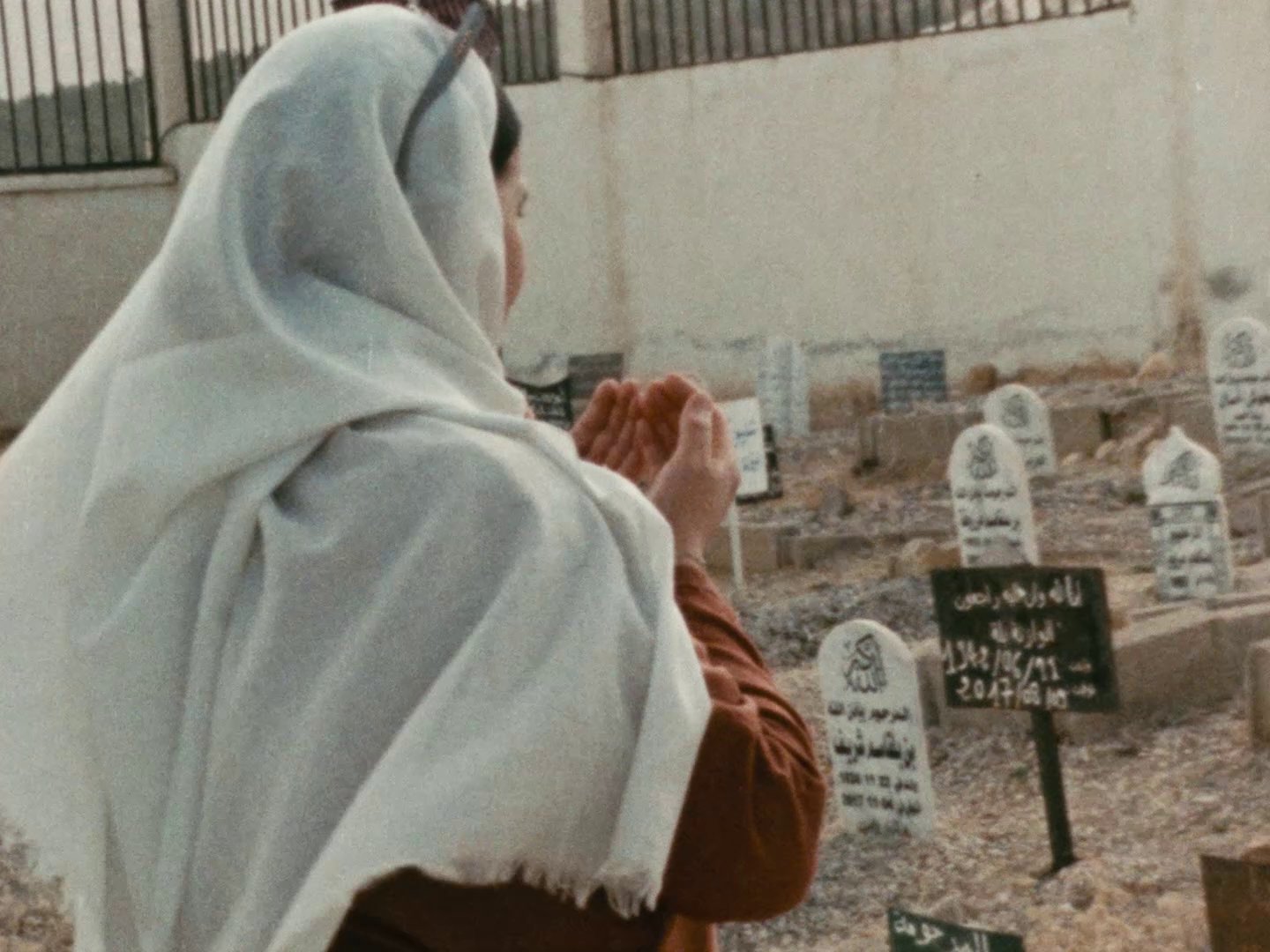 Woman wearing a head scarf facing away from the camera looking at gravestones in the background