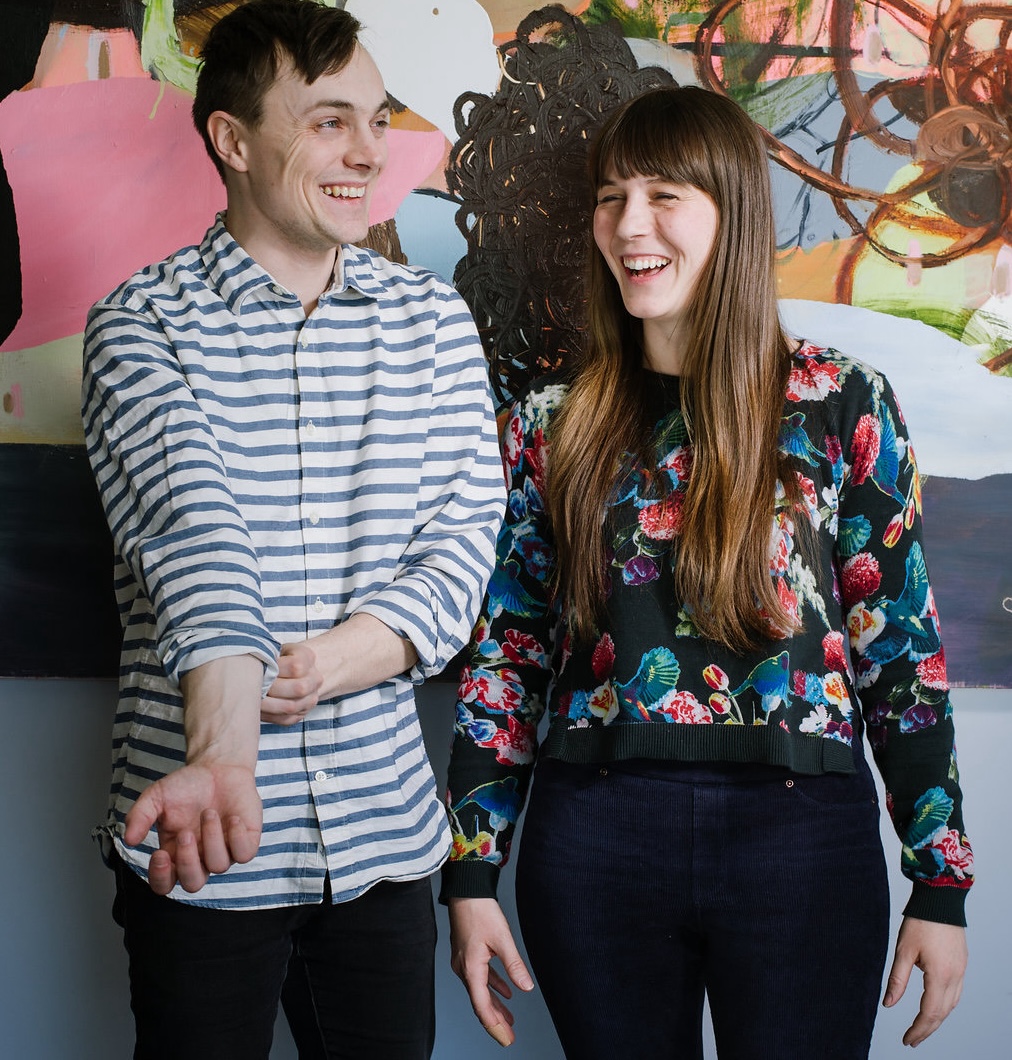 Two people standing in front of their artwork. The man on the left is wearing a striped shirt, the woman on the right is wearing a floral top. Both are smiling and not looking at the camera.