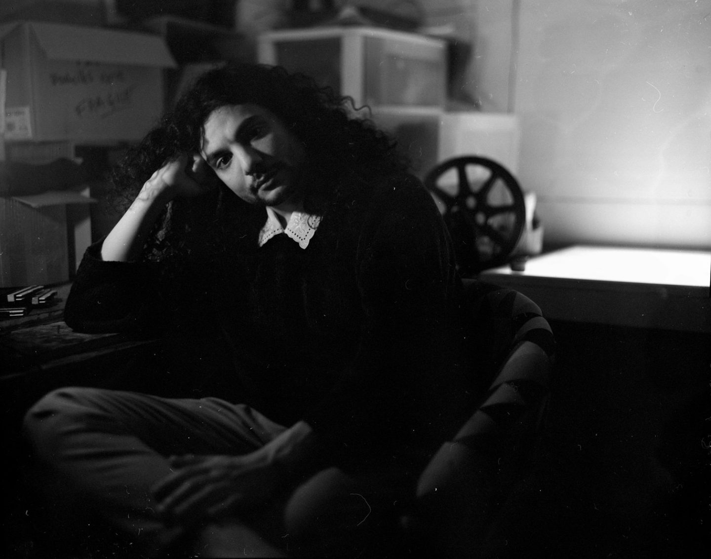 Shadowy portrait of a person with long dark hair sitting at a table with their fist on their temple.