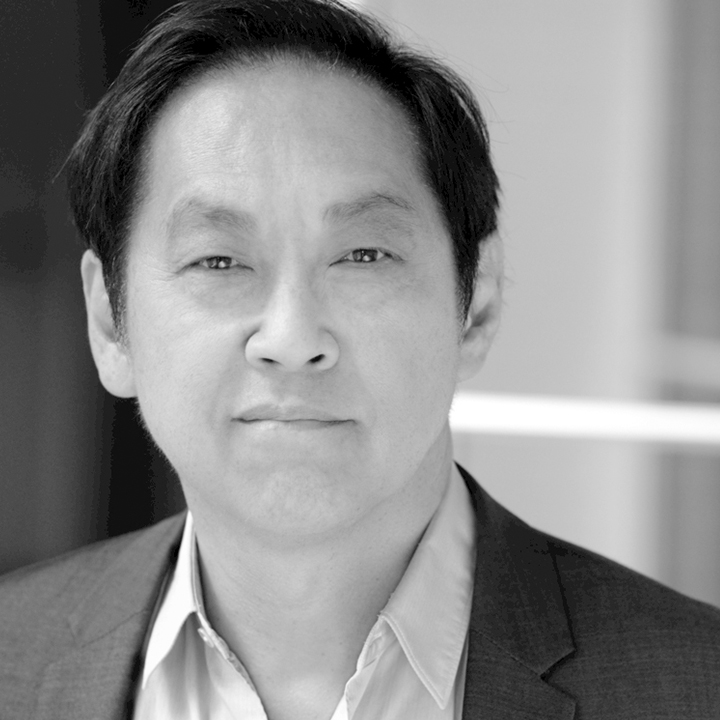 Black and white image of a man looking at camera. He is wearing a white dress shirt and dark jacket.