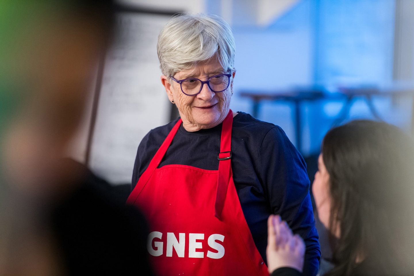 Woman with short grey hair wearing a red apron. The apron reads "Agnes"