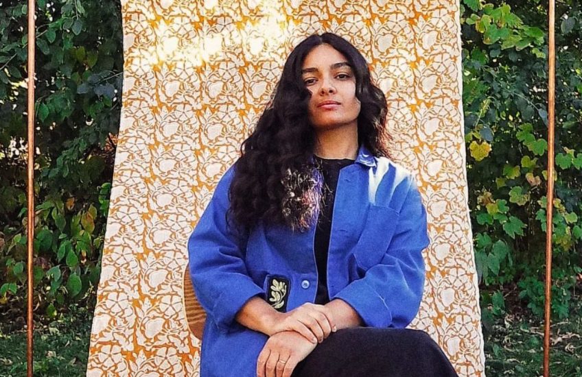 A woman with long wavy hair and a blue coat sits on a chair outside. Behind her a dyed tapestry is hung infront of foliage.
