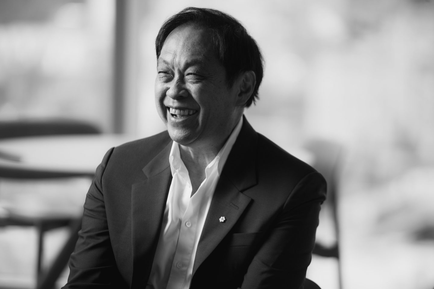 Black and white photograph of a man laughing. He is seen in three quarter profile and is wearing a suit jacket and dress shirt, with the top button undone.