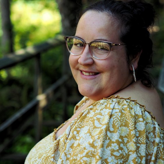 white woman wearing yellow floral print dress and glasses. She is looking over her left shoulder at the camera and smiling.