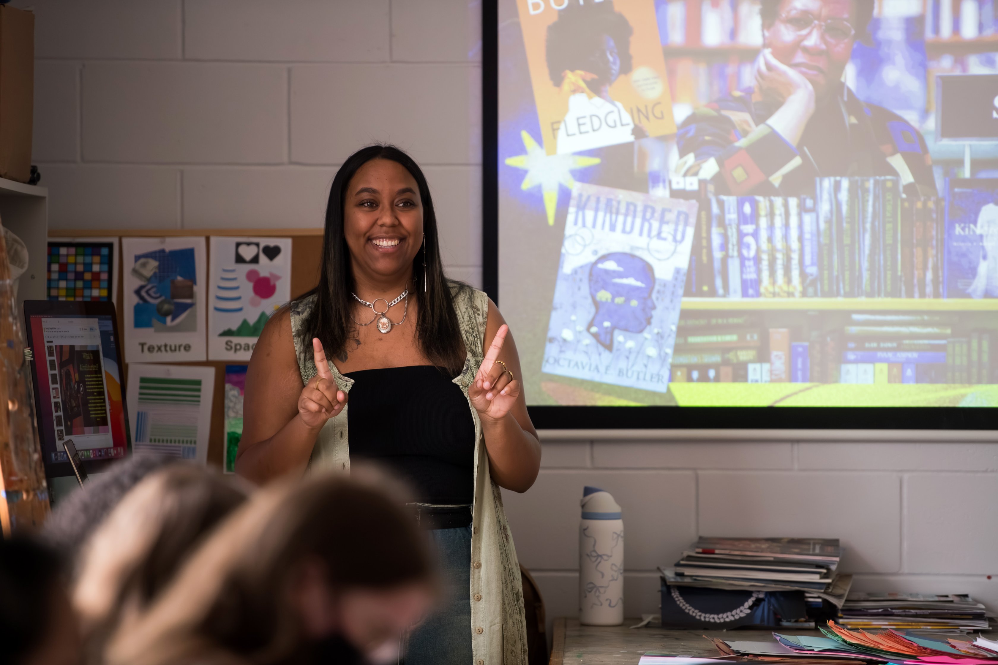 Young Black woman standing at the front of a classroom with a projection behind her. She is smiling and looks welcoming.